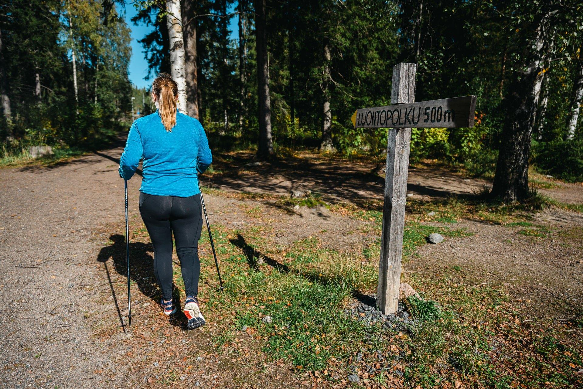 Sauvakävelevä nainen kävelee selin kameraan. Taustalla on metsää ja naisen vieressä on kyltti, jossa lukee Luontopolku 500 m.