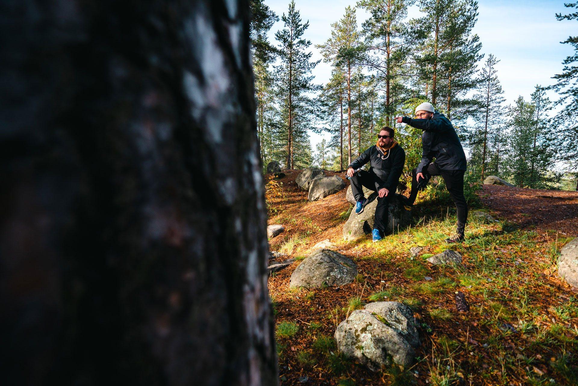 Kuvassa on kaksi miestä, joista toinen istuu kivellä ja toinen seisoo hänen vieressään ja osoittaa kädellä eteensä. Taustalla on metsää.