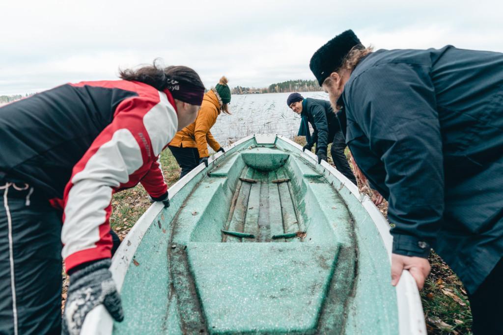 Neljä henkilöä työntää venettä vesille syksyisessä järvenrantamaisemassa.