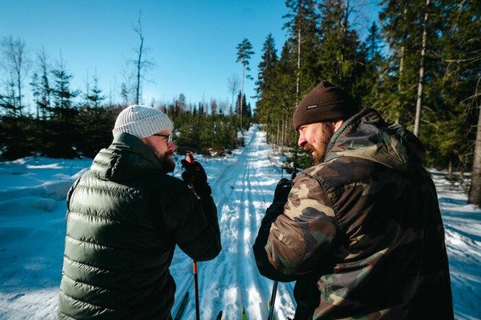 Kuvassa kaksi miestä nojailee hiihtosauvoihin ja katsoo toisiinsa. Taustalla on talvinen metsämaisema.