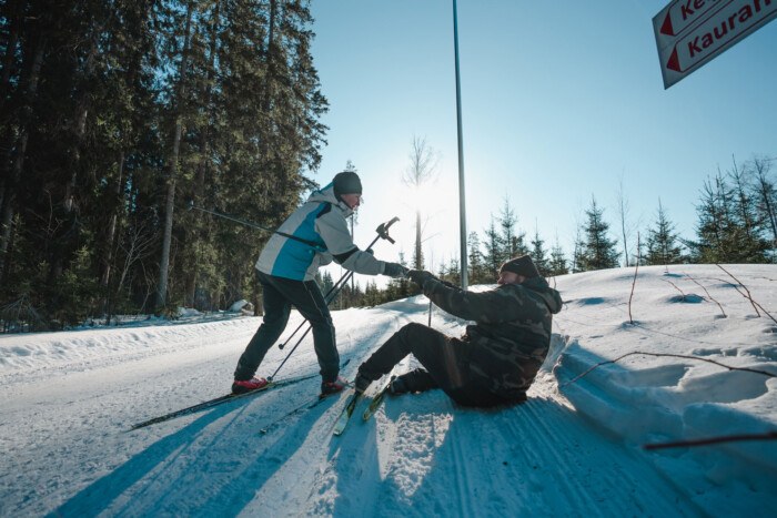 Kuvassa nainen auttaa maahan kaatunutta miestä ylös hiihtoladulla. Molemmilla on sukset jalassa ja sauvat käsissä. Maisema on talvinen ja taustalla paistaa aurinko.