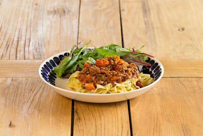 Kuvassa on puupöydän päällä oleva lautanen, jossa on spagettia, jauhelihakastiketta ja salaattia.