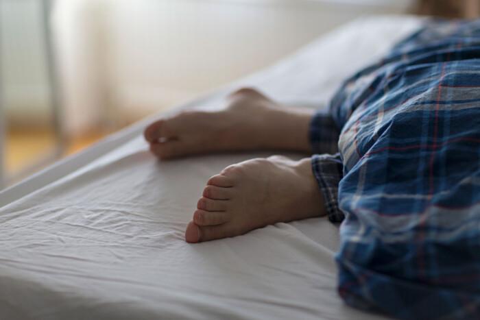 Kuvassa näkyvät siniruudullisen peiton alta pilkottavat paljaat jalat sängyllä.
