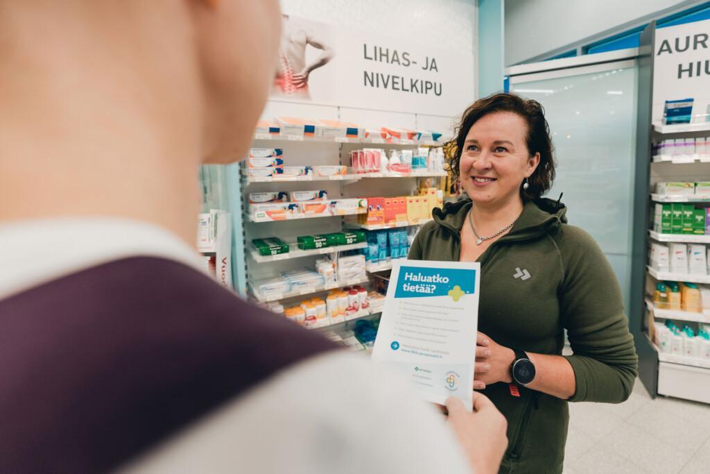 Kuvassa nainen kameraan päin ja toisen henkilön hahmo etualalla selin kameraan. Etualalla olevalla henkilöllä paperi kädessään. Taustalla näkyy apteekin myymäläkalustoa.