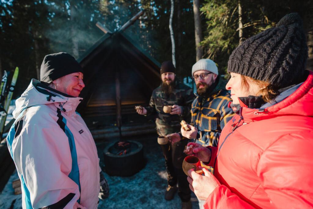 Kuvassa neljä henkilöä seisoo talvivaatteet päällä puisen laavun edustalla kuksat käsissään.