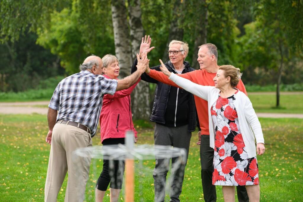 Kuvassa viisi ikäihmistä lyö käsiä yhteen frisbeegolf-korin edessä. Taustalla näkyy vihreää puistomaista maisemaa.